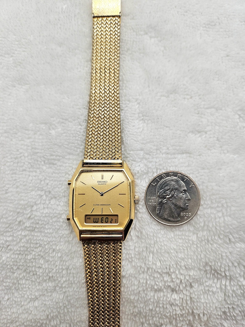 May include: A gold-toned Seiko Quartz alarm chronograph watch with a mesh band. The watch face is rectangular with a digital display showing the date. A US quarter is next to the watch for size comparison.
