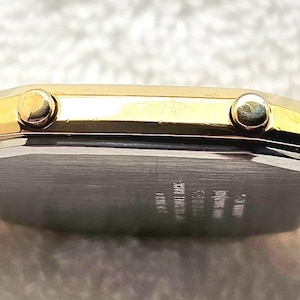 May include: A gold-toned rectangular wristwatch with a light brown fabric band. The watch face is not visible, but the side profile shows two small buttons and a silver-colored back. The band appears to be woven.