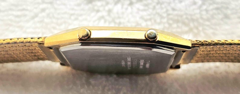 May include: A gold-toned rectangular wristwatch with a light brown fabric band. The watch face is not visible, but the side profile shows two small buttons and a silver-colored back. The band appears to be woven.