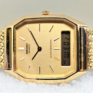May include: A gold-toned Seiko Quartz watch with a rectangular face and a woven gold band. The watch face displays the time and day, with the text "SEIKO QUARTZ" visible. The watch has a digital display showing the date.