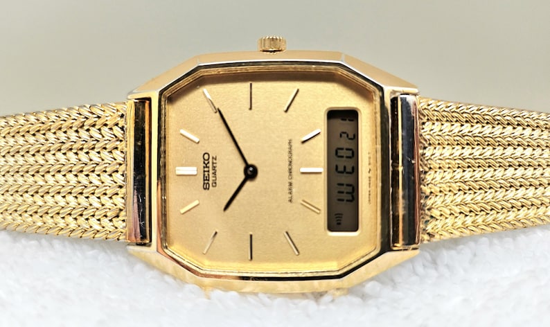May include: A gold-toned Seiko Quartz watch with a rectangular face and a woven gold band. The watch face displays the time and day, with the text "SEIKO QUARTZ" visible. The watch has a digital display showing the date.