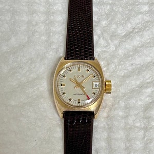 May include: A gold-toned Elgin automatic wristwatch with a white face and black leather band. The watch face has black hour and minute hands, a red second hand, and a small date window at the 3 o'clock position.