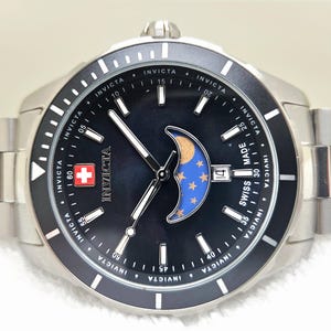 Puede incluir: Un reloj Invicta de acero inoxidable con esfera negra, que presenta un dise&ntilde;o de fase lunar con detalles dorados y azules. El reloj tiene una pantalla de fecha y un emblema de la cruz suiza. La correa del reloj tambi&eacute;n es de acero inoxidable.