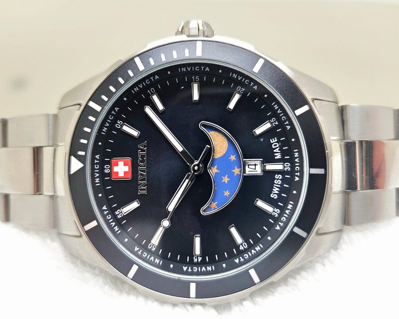Puede incluir: Un reloj Invicta de acero inoxidable con esfera negra, que presenta un dise&ntilde;o de fase lunar con detalles dorados y azules. El reloj tiene una pantalla de fecha y un emblema de la cruz suiza. La correa del reloj tambi&eacute;n es de acero inoxidable.