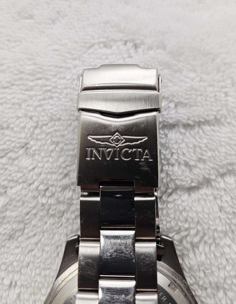 Puede incluir: Primer plano de un cierre de reloj Invicta plateado. El cierre es rectangular con el nombre de la marca y el logotipo grabados en la parte delantera. La correa del reloj tambi&eacute;n es plateada y parece ser de metal.