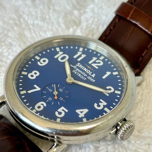 Może przedstawiać: Zbliżenie zegarka Shinola Argonite-1069 Detroit z niebieską tarczą i białymi cyframi. Zegarek ma srebrną kopertę i brązowy sk&oacute;rzany pasek. Tarcza zegarka zawiera małą tarczę pomocniczą, a godzina to około 10:10.