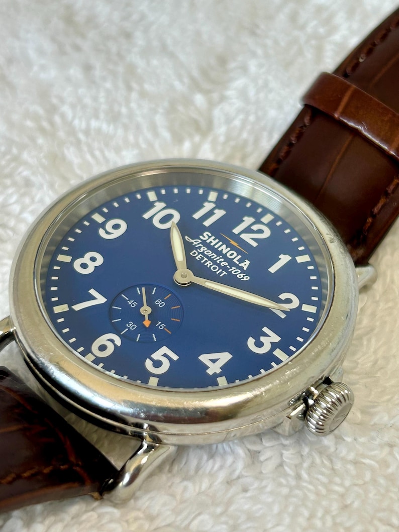 Może przedstawiać: Zbliżenie zegarka Shinola Argonite-1069 Detroit z niebieską tarczą i białymi cyframi. Zegarek ma srebrną kopertę i brązowy sk&oacute;rzany pasek. Tarcza zegarka zawiera małą tarczę pomocniczą, a godzina to około 10:10.