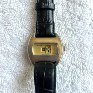 May include: A vintage digital watch with a rectangular gold-toned face displaying the time as 10:10. The watch has a silver-colored case and a black leather band. The watch face has the word "DIGITAL" printed on it.