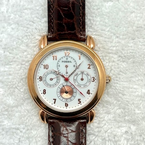 Könnte beinhalten: Eine Fossil Armbanduhr mit weißem Zifferblatt, goldfarbenem Gehäuse und braunem Lederarmband. Die Uhr verfügt über mehrere Hilfszifferblätter, darunter Tages-, Datums- und Monatsanzeigen. Klassisches Design mit römischen Ziffern und roten Zeigern.