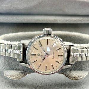 Pode incluir: Um relógio Omega Genève vintage com caixa prateada e mostrador claro. O relógio apresenta ponteiros e marcadores de horas prateados e uma pulseira prateada. O relógio é exibido em uma superfície cinza.