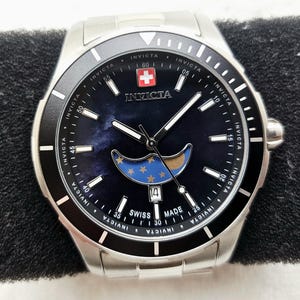 Puede incluir: Un reloj Invicta plateado con una esfera azul oscuro con un dise&ntilde;o de luna y estrellas. El reloj tiene un bisel negro y una pantalla de fecha en la posici&oacute;n de las 4 en punto. La esfera del reloj tambi&eacute;n tiene la bandera suiza y la inscripci&oacute;n "Swiss Made".
