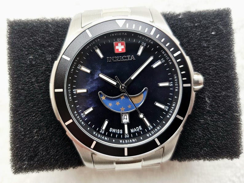 Puede incluir: Un reloj Invicta plateado con una esfera azul oscuro con un dise&ntilde;o de luna y estrellas. El reloj tiene un bisel negro y una pantalla de fecha en la posici&oacute;n de las 4 en punto. La esfera del reloj tambi&eacute;n tiene la bandera suiza y la inscripci&oacute;n "Swiss Made".