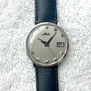 Puede incluir: Un reloj Mido Ocean Star vintage con esfera plateada, visualización de fecha y agujas negras. El reloj tiene una caja de acero inoxidable y una correa de cuero azul oscuro. La esfera del reloj tiene el texto "Mido Ocean Star" y "Powerwind".