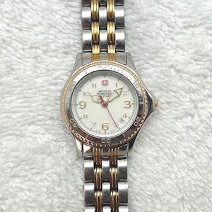 May include: A Wenger Swiss watch with a silver and gold-tone stainless steel band and a white dial. The watch face features black numerals, gold-tone hands, and a red second hand. The watch has a gold-tone bezel.