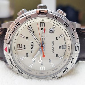 May include: Silver-toned Timex Compass watch with a light-colored face, black leather band, and red accents. The watch features a compass bezel and a date display. The watch face displays the time and the word "COMPASS".