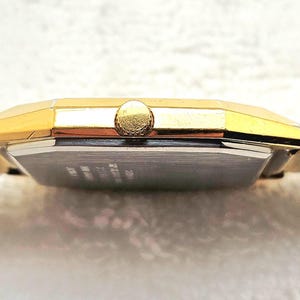 May include: A gold-toned wristwatch with a rectangular face and a textured band. The watch has a gold crown and a silver-colored face. The band appears to be made of a woven material. The watch is displayed on a white surface.