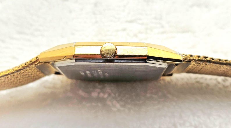 May include: A gold-toned wristwatch with a rectangular face and a textured band. The watch has a gold crown and a silver-colored face. The band appears to be made of a woven material. The watch is displayed on a white surface.