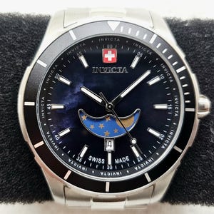 Puede incluir: Reloj Invicta de acero inoxidable con bisel negro y esfera azul oscuro. La esfera del reloj presenta un dise&ntilde;o de fase lunar con estrellas y una visualizaci&oacute;n de la fecha. El reloj tiene una etiqueta Swiss Made y el logotipo de Invicta.
