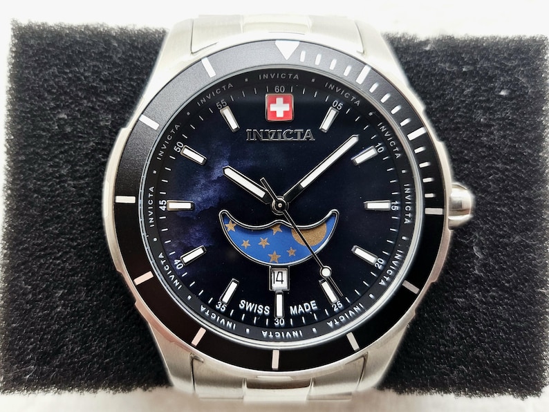 Puede incluir: Reloj Invicta de acero inoxidable con bisel negro y esfera azul oscuro. La esfera del reloj presenta un dise&ntilde;o de fase lunar con estrellas y una visualizaci&oacute;n de la fecha. El reloj tiene una etiqueta Swiss Made y el logotipo de Invicta.