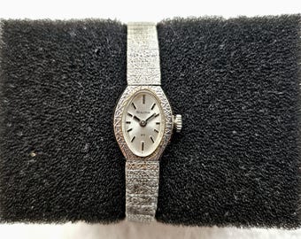 Könnte beinhalten: Eine Vintage-Bulova-Armbanduhr mit silberfarbenem, ovalem Zifferblatt und strukturiertem Silberarmband. Das Zifferblatt hat schwarze Stundenmarkierungen und Zeiger. Die Uhr wird auf einer schwarzen, strukturierten Oberfläche präsentiert.