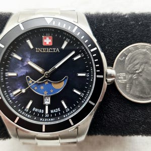 Puede incluir: Un reloj Invicta de acero inoxidable con bisel negro y esfera azul oscuro con dise&ntilde;o de fase lunar. El reloj tiene una pantalla de fecha y est&aacute; junto a una moneda.