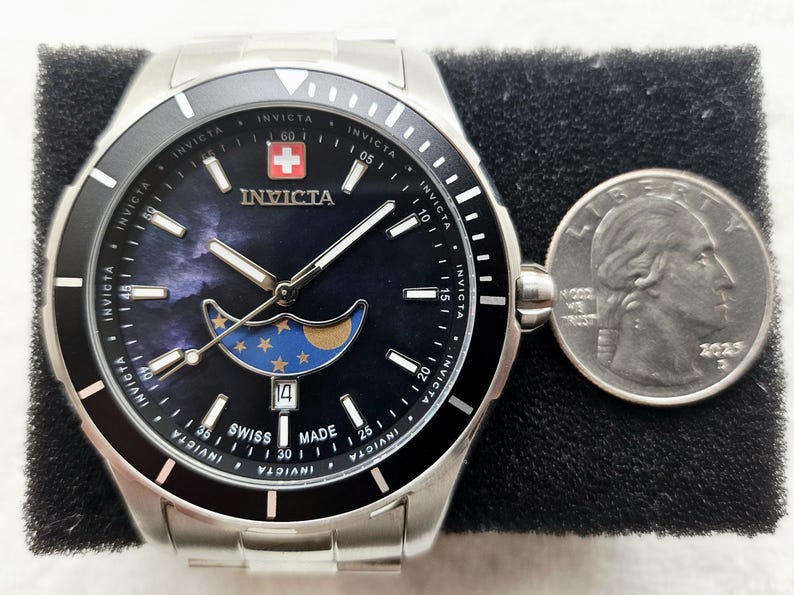 Puede incluir: Un reloj Invicta de acero inoxidable con bisel negro y esfera azul oscuro con dise&ntilde;o de fase lunar. El reloj tiene una pantalla de fecha y est&aacute; junto a una moneda.