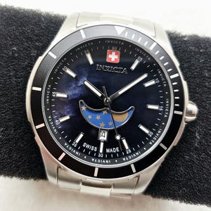 Puede incluir: Un reloj Invicta de acero inoxidable con una esfera azul oscuro con un dise&ntilde;o de luna y estrellas. El reloj tiene un bisel negro, agujas blancas y una pantalla de fecha. El reloj est&aacute; marcado "Swiss Made".