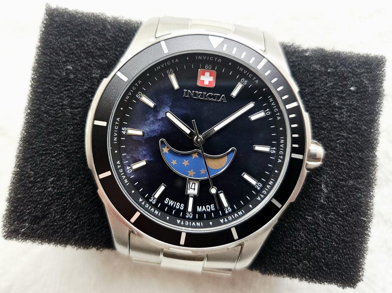 Puede incluir: Un reloj Invicta de acero inoxidable con una esfera azul oscuro con un dise&ntilde;o de luna y estrellas. El reloj tiene un bisel negro, agujas blancas y una pantalla de fecha. El reloj est&aacute; marcado "Swiss Made".