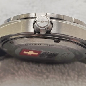 Puede incluir: Primer plano de un reloj de pulsera plateado con una correa de metal. La esfera del reloj no es visible, pero la corona y la parte trasera del reloj est&aacute;n a la vista. La parte trasera del reloj tiene el texto "WATER RESISTANT 100M" y un emblema rojo y blanco.
