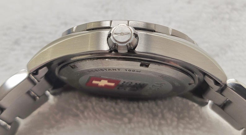 Puede incluir: Primer plano de un reloj de pulsera plateado con una correa de metal. La esfera del reloj no es visible, pero la corona y la parte trasera del reloj est&aacute;n a la vista. La parte trasera del reloj tiene el texto "WATER RESISTANT 100M" y un emblema rojo y blanco.