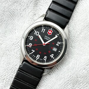 May include: A Swiss Army wristwatch with a black dial, white Arabic numerals, a red second hand, and a date window at the three o'clock position. The watch features a silver-tone stainless steel case and a black rubber strap with horizontal ridges.