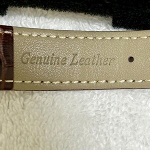 Może przedstawiać: Zbliżenie paska do zegarka z wytłoczonymi słowami "Genuine Leather" na jasnobeżowej spodniej stronie. Pasek jest brązowy z białymi przeszyciami wzdłuż krawędzi.