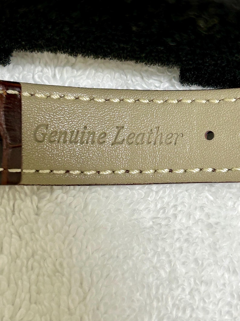 Może przedstawiać: Zbliżenie paska do zegarka z wytłoczonymi słowami "Genuine Leather" na jasnobeżowej spodniej stronie. Pasek jest brązowy z białymi przeszyciami wzdłuż krawędzi.