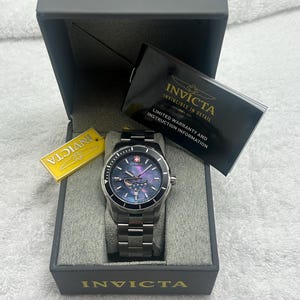 Puede incluir: Un reloj Invicta plateado con esfera azul y correa de acero inoxidable se muestra en una caja gris. El reloj tiene un bisel negro y una etiqueta que dice "Invicta". La caja tambi&eacute;n contiene una tarjeta de garant&iacute;a.