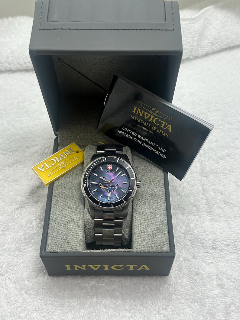 Puede incluir: Un reloj Invicta plateado con esfera azul y correa de acero inoxidable se muestra en una caja gris. El reloj tiene un bisel negro y una etiqueta que dice "Invicta". La caja tambi&eacute;n contiene una tarjeta de garant&iacute;a.