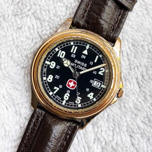 May include: A Swiss Military wristwatch with a round gold-tone case, a black dial featuring white Arabic numerals, a date window at the three o'clock position, a red and white cross logo, and a dark brown textured leather strap with a buckle.