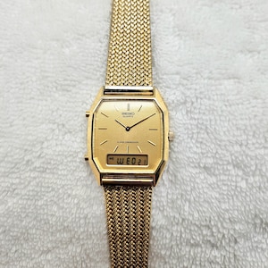 May include: A gold-toned Seiko Quartz alarm chronograph watch with a mesh band. The watch face is rectangular with a digital display showing the date. The watch hands and hour markers are black. The band is a woven mesh design.