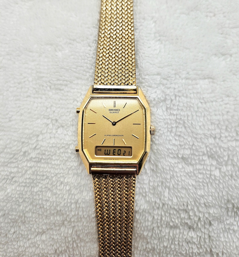 May include: A gold-toned Seiko Quartz alarm chronograph watch with a mesh band. The watch face is rectangular with a digital display showing the date. The watch hands and hour markers are black. The band is a woven mesh design.
