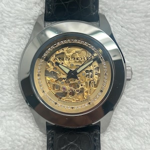 May include: Silver Android automatic watch with a gold skeleton dial and a black leather strap. The watch face is encircled by small, clear stones. The word "ANDROID" is visible on the watch face.