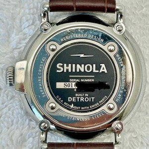 Może przedstawiać: Zbliżenie zegarka Shinola z brązowym sk&oacute;rzanym paskiem. Tarcza zegarka jest czarna z białym napisem "SHINOLA" i "DETROIT". Zegarek jest ze stali nierdzewnej i ma szafirowe szkło.