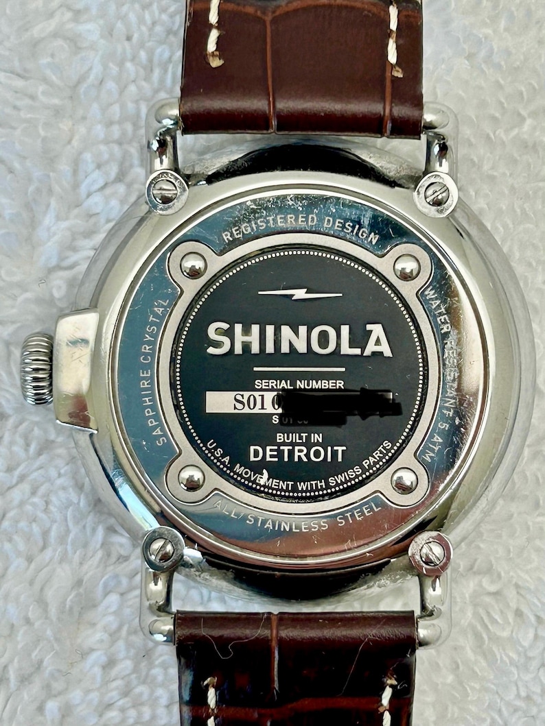 Może przedstawiać: Zbliżenie zegarka Shinola z brązowym sk&oacute;rzanym paskiem. Tarcza zegarka jest czarna z białym napisem "SHINOLA" i "DETROIT". Zegarek jest ze stali nierdzewnej i ma szafirowe szkło.