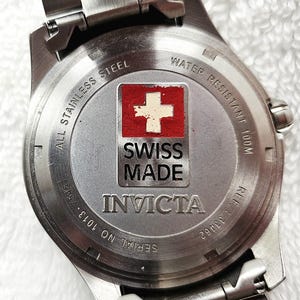 Puede incluir: Primer plano de la parte trasera de un reloj Invicta de acero inoxidable. La esfera del reloj presenta la bandera suiza y las palabras "SWISS MADE". El reloj es resistente al agua hasta 100m. N&uacute;mero de serie 1013-75451 y referencia 33462 visibles.