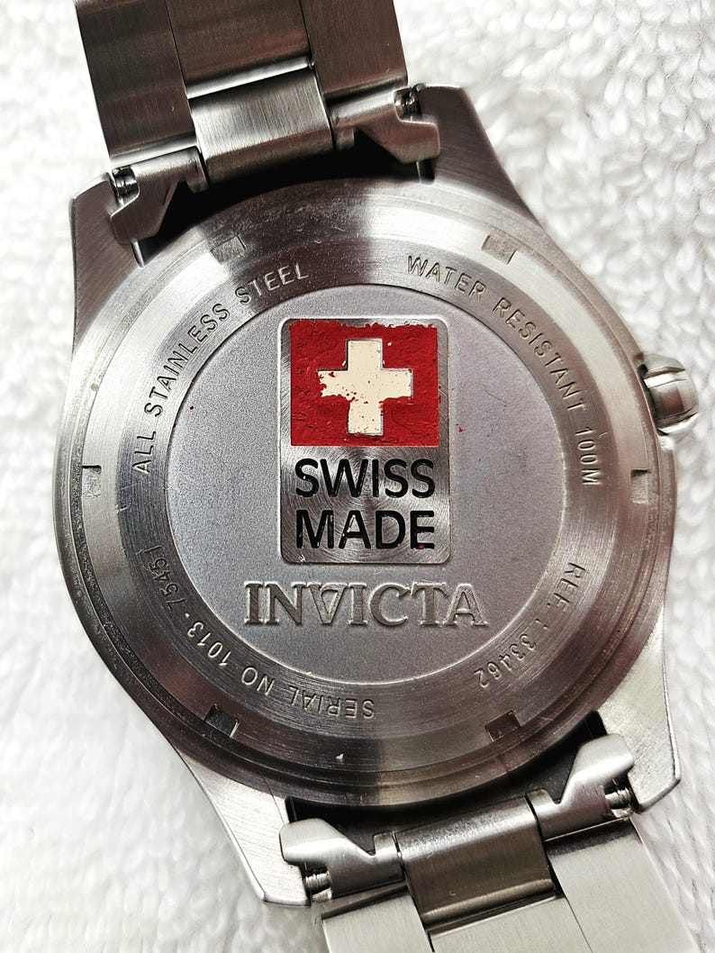 Puede incluir: Primer plano de la parte trasera de un reloj Invicta de acero inoxidable. La esfera del reloj presenta la bandera suiza y las palabras "SWISS MADE". El reloj es resistente al agua hasta 100m. N&uacute;mero de serie 1013-75451 y referencia 33462 visibles.