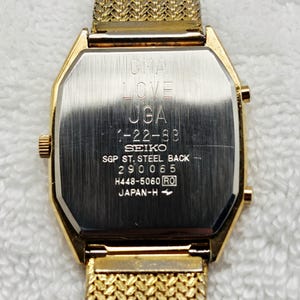 May include: Gold-toned Seiko watch with a mesh band. The watch face is rectangular with rounded edges. The back of the watch has engraved text including "OMA LOVE UGA 1-22-88 SEIKO SGP ST. STEEL BACK 290065 H448-5060 RO JAPAN-H".