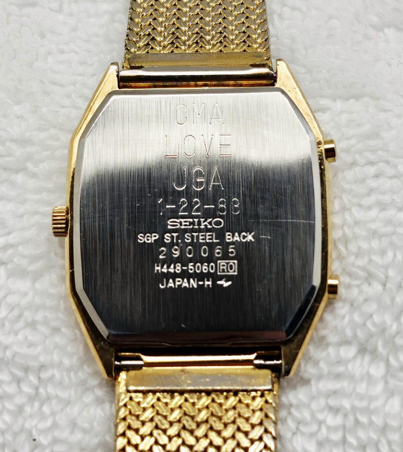 May include: Gold-toned Seiko watch with a mesh band. The watch face is rectangular with rounded edges. The back of the watch has engraved text including "OMA LOVE UGA 1-22-88 SEIKO SGP ST. STEEL BACK 290065 H448-5060 RO JAPAN-H".