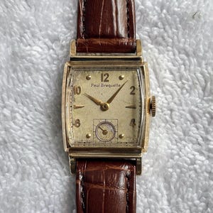 May include: A vintage gold-toned Paul Breguette wristwatch with a rectangular face and a brown leather band. The watch face displays the numbers 1 through 12, with the brand name printed below the 12. The watch has a small seconds dial.