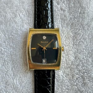 Puede incluir: Un reloj Bulova vintage con una caja rectangular dorada y una esfera negra. El reloj presenta agujas doradas, un pequeño acento de diamante y una ventana de fecha. Está sujeto a una correa de cuero negro.