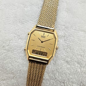 May include: A gold-toned Seiko Quartz watch with a mesh band. The watch face is rectangular with a digital display showing the date. The watch has a gold-colored bezel and hands. The band is a woven mesh design.