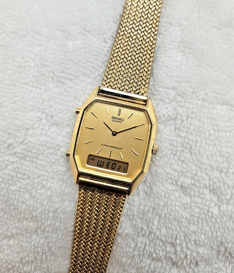 May include: A gold-toned Seiko Quartz watch with a mesh band. The watch face is rectangular with a digital display showing the date. The watch has a gold-colored bezel and hands. The band is a woven mesh design.