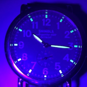 Może przedstawiać: Zbliżenie tarczy zegarka Shinola Argonite-1069 podświetlonej niebieskim światłem. Zegarek ma świecące wskaz&oacute;wki i znaczniki, z widocznym tekstem "SHINOLA Argonite-1069 DETROIT". Tarcza zegarka wyświetla liczby od 1 do 12.